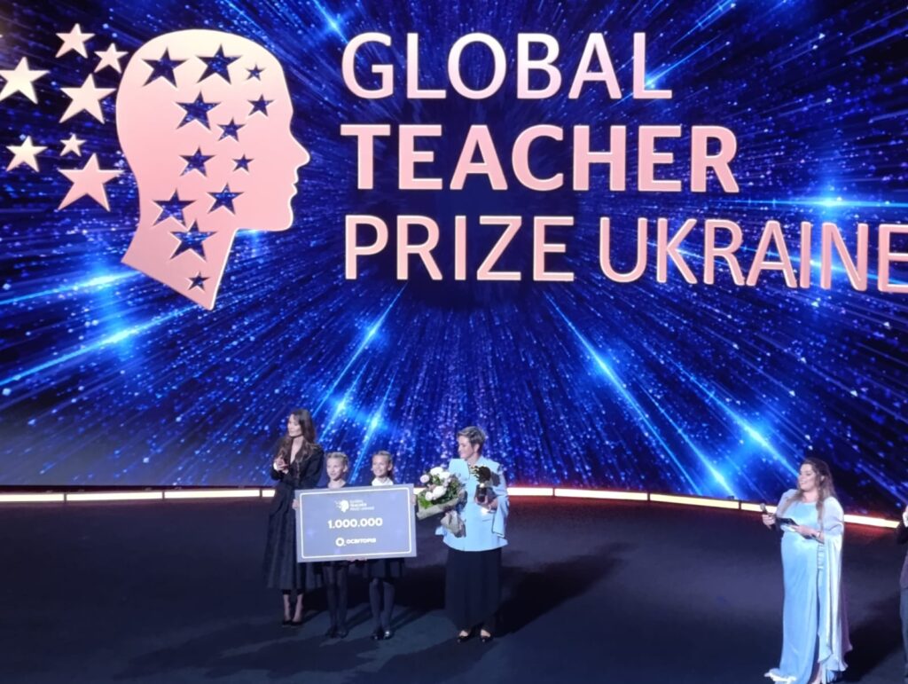 Сцена премії Global Teacher Prize Ukraine 2026, де на тлі великого синього екрана з логотипом стоять ведучі та переможниця Світлана Меркулова, тримаючи в руках квіти, нагороду та символічний чек на 1 000 000 гривень.